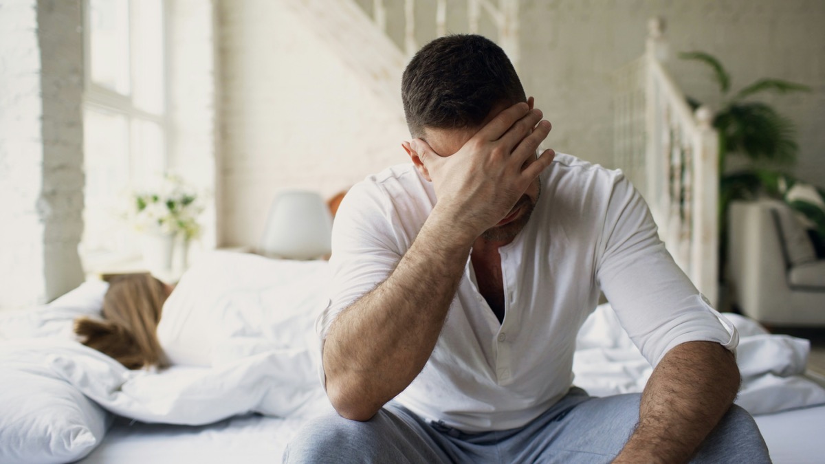 A stressed man sitting on a bed with his hand over his face.